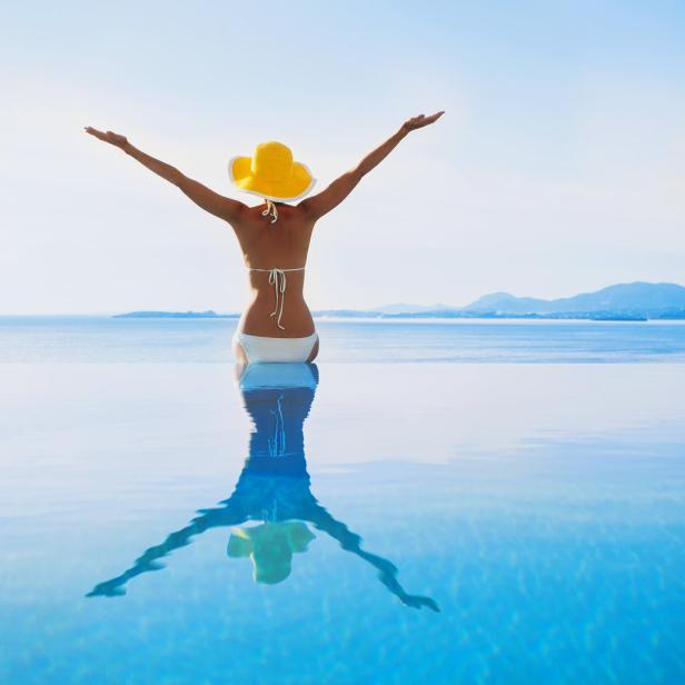 Eine Frau mit gelbem Hut sitzt am Rand eines Infinity-Pools und streckt die Arme in die Luft.