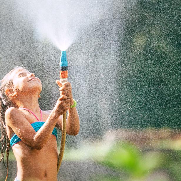 Ein süßes kleines Mädchen übergießt sich mit einem Wasserschlauch und erzeugt so einen Regen. Sommerliche Freude.