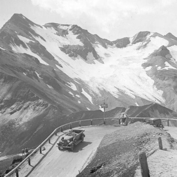 Ein Auto fährt auf einer kurvenreichen Straße vor einer Bergkulisse mit Schnee.