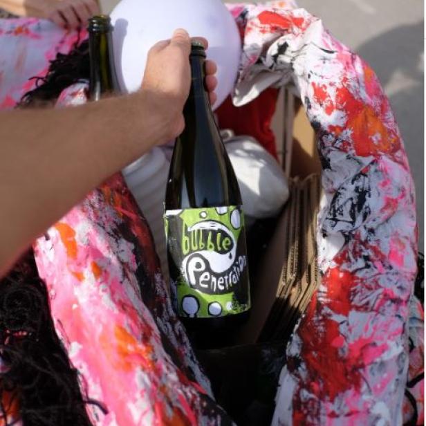 Eine Hand hält eine Flasche „Bubble Penetration“ in einem Karton mit einer bemalten Skulptur.