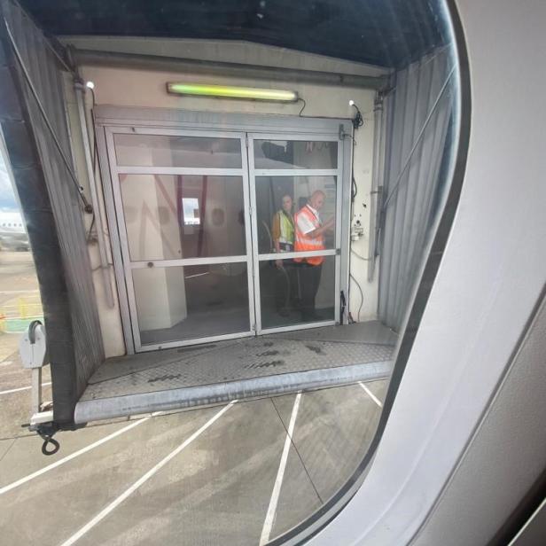 Blick aus einem Flugzeugfenster auf eine Fluggastbrücke mit zwei Flughafenmitarbeitern.