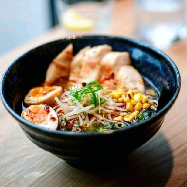 Eine Schüssel Ramen mit Hühnchen, Ei, Mais und Gemüse steht auf einem Holztisch.