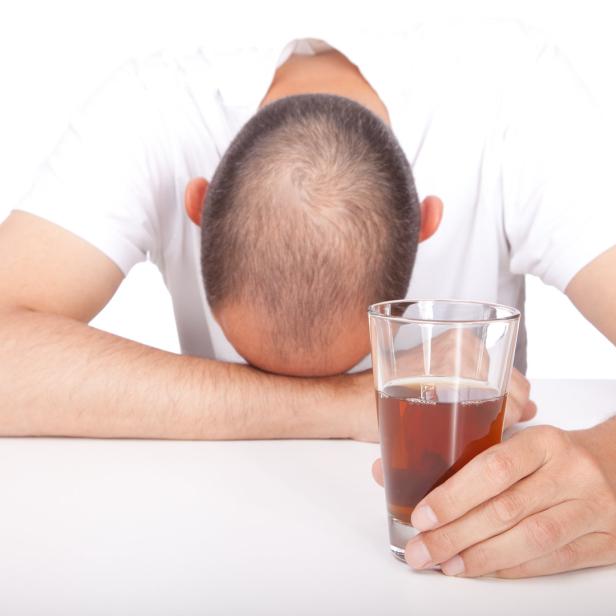 Ein Mann liegt erschöpft auf dem Tisch, ein Glas mit einem Getränk in der Hand.