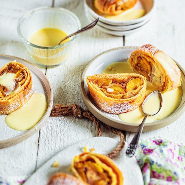 Mehrere Teller mit Apfelstrudel und Vanillesauce auf einem weißen Holztisch.