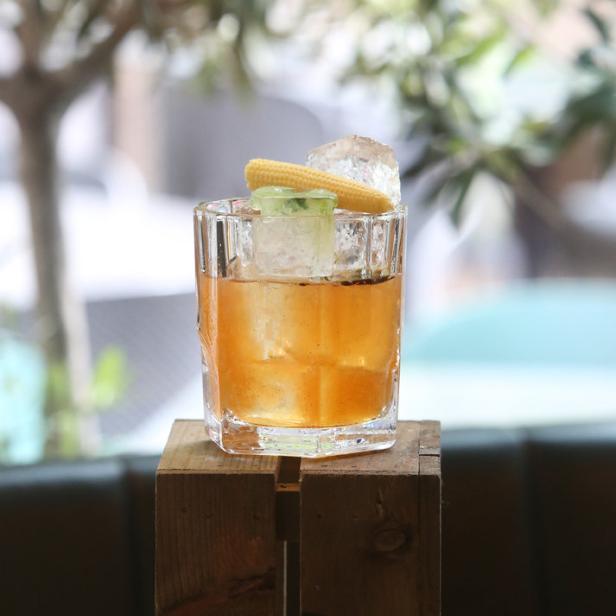 Ein Cocktail mit Eiswürfeln und einem Stück Zuckermais als Dekoration steht auf einer Holzkiste.