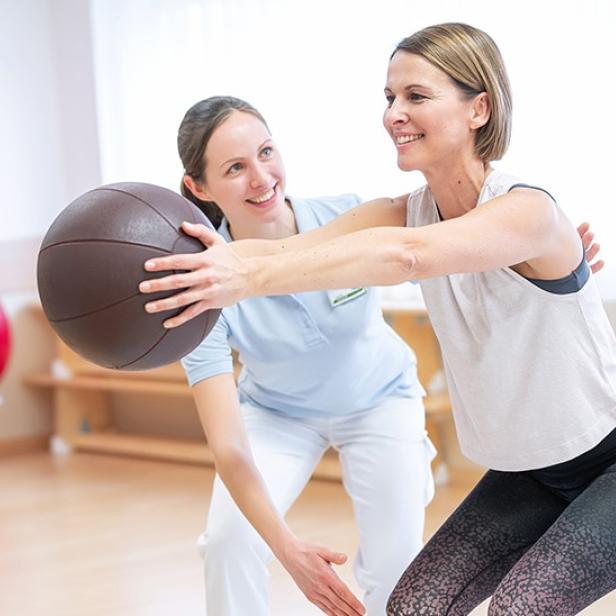 Eine Frau macht mit einem Medizinball Kniebeugen unter Aufsicht einer Physiotherapeutin.