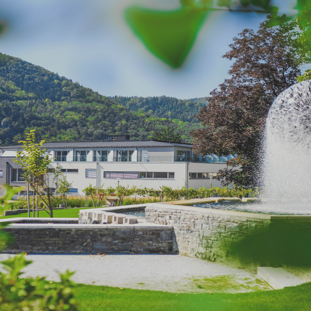 Ein kugelförmiger Springbrunnen sprüht vor einem modernen Gebäude und einem bewaldeten Hügel.