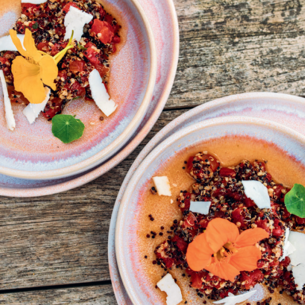 Zwei Teller mit Quinoa-Salat, Tomaten, Kokosraspeln und essbaren Blüten auf einem Holztisch.