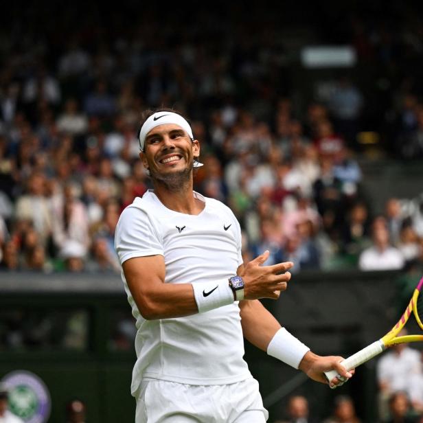 Rafael Nadal lächelt auf einem Tennisplatz mit Zuschauern im Hintergrund.