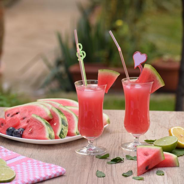 Zwei Gläser Wassermelonensaft stehen auf einem Holztisch, dekoriert mit Limetten und Melonenstücken.
