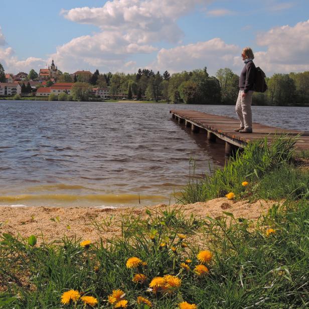 Eine Frau steht auf einem Holzsteg an einem See, im Hintergrund eine Ortschaft.