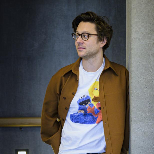 Ein Mann mit Brille, brauner Jacke und einem T-Shirt mit Krümelmonster-Motiv lehnt an einer Wand.