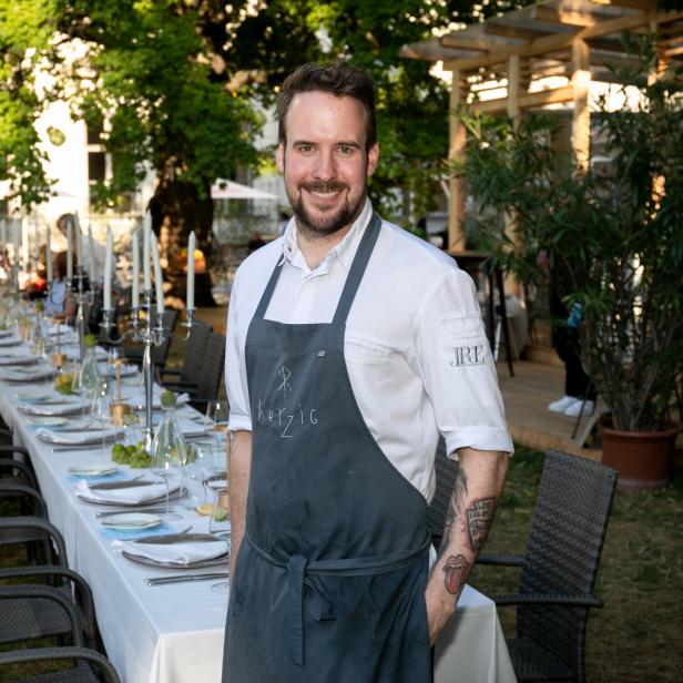 Ein Koch steht vor einer langen, festlich gedeckten Tafel im Freien.
