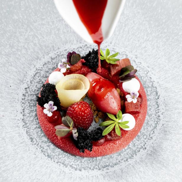 Eine rote Sauce wird über ein kunstvolles Dessert mit Himbeeren und Blüten gegossen.