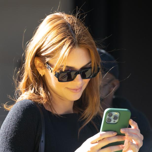 Eine Frau mit Sonnenbrille und roten Haaren schaut auf ihr Smartphone.