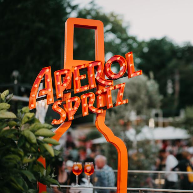 Ein beleuchtetes „Aperol Spritz“-Schild steht vor einer belebten Bar im Freien.