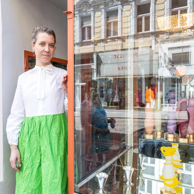 Eine Frau in einem weißen Oberteil und einem grünen Rock steht in der Tür eines Ladens.