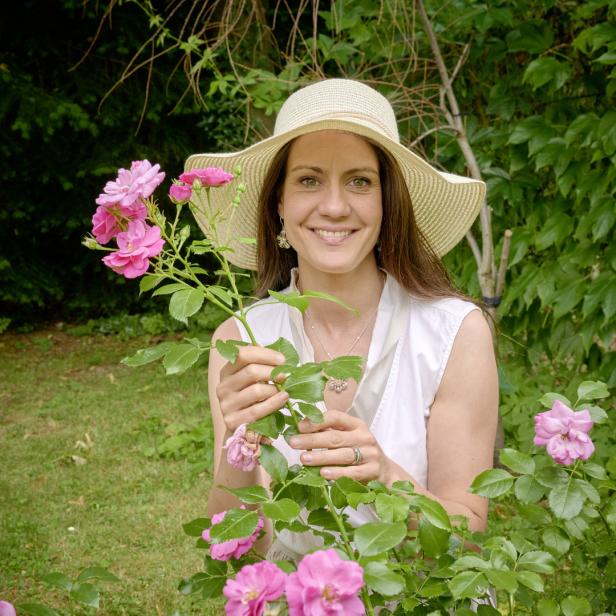 Eine lächelnde Frau mit Hut betrachtet Rosen in einem Garten.