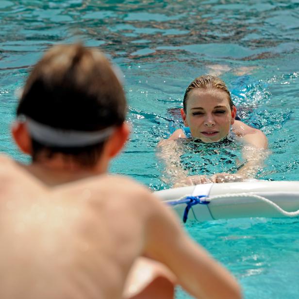 Eine Frau im Schwimmbad hält sich an einem Rettungsring fest, während ein Junge zusieht.