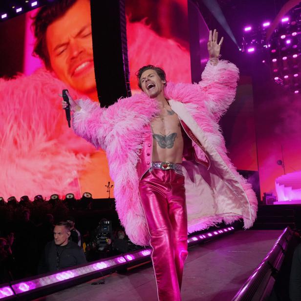 Harry Styles tritt in einem rosa Federboa-Mantel und einer glänzenden Hose auf einer Bühne auf.