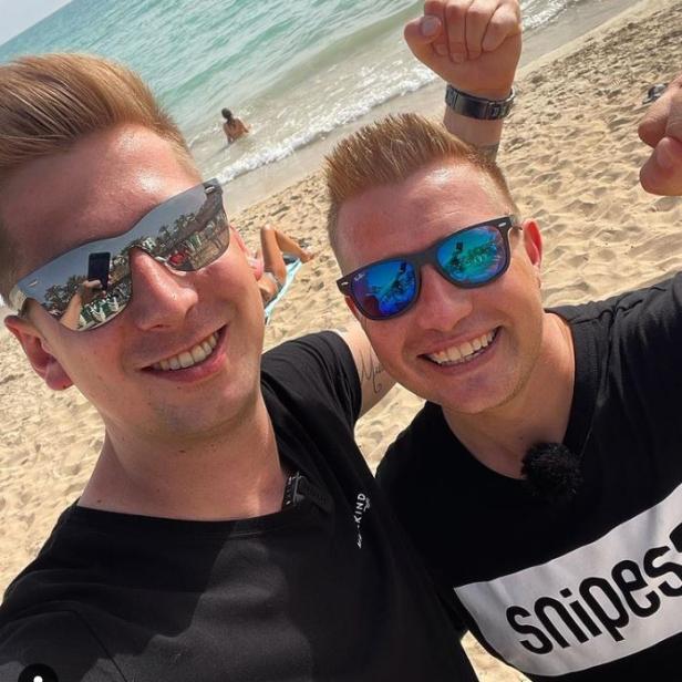 Zwei Männer mit Sonnenbrillen posieren am Strand vor dem Meer.