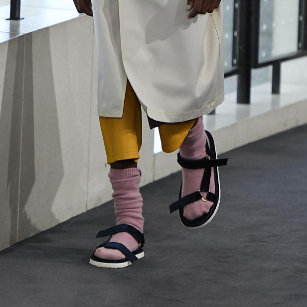 Eine Person trägt Socken in Sandalen und gelbe Shorts unter einem weißen Rock.