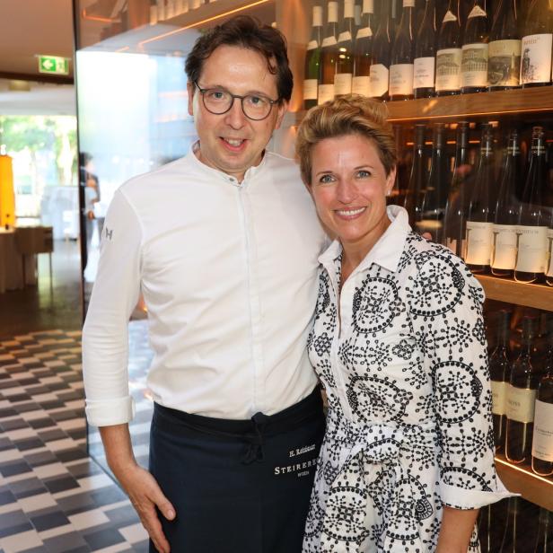 Ein Mann in Kochkleidung und eine Frau stehen vor einem Regal mit Weinflaschen.