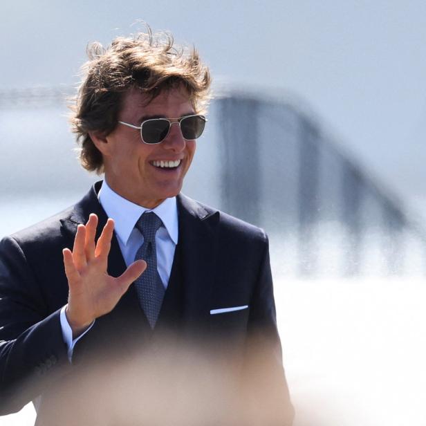 Tom Cruise in Anzug und Sonnenbrille winkt lächelnd.