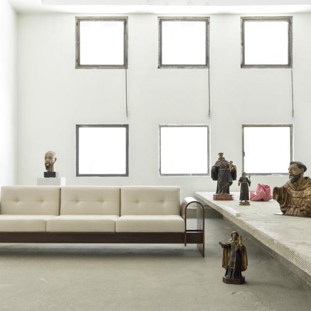 Ein beiges Sofa steht in einem Raum mit Skulpturen und sechs Fenstern.