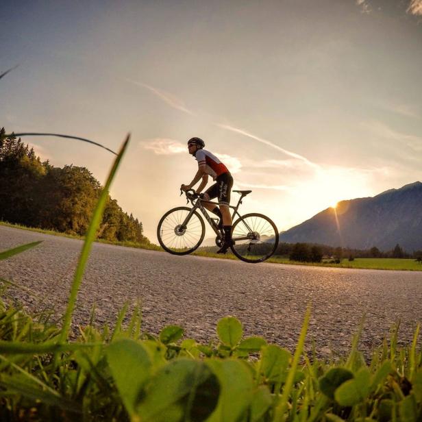 Ein Radfahrer fährt auf einer Landstraße vor einer Bergkulisse in Richtung Sonnenuntergang.