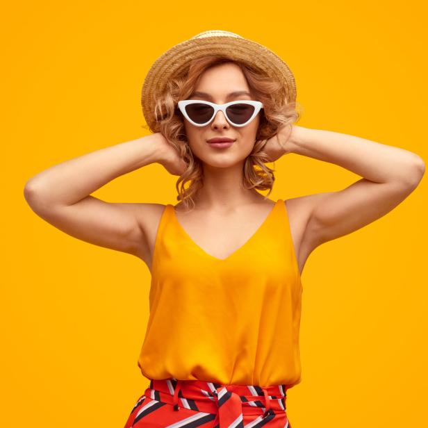 Eine Frau mit Sonnenbrille und Hut posiert vor einem orangefarbenen Hintergrund.