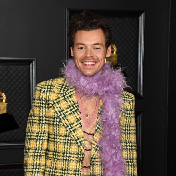 Harry Styles in einem gelb-karierten Sakko und einer lila Federboa lächelt.
