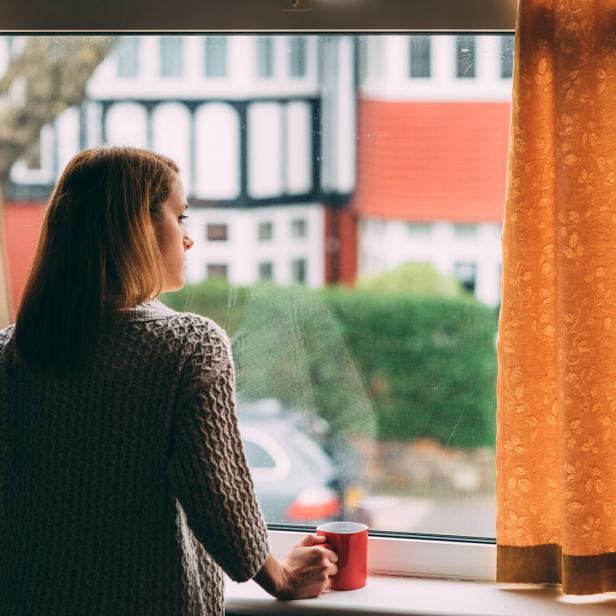 Eine Frau steht mit einer roten Tasse in der Hand am Fenster und blickt nach draußen zwischen gelben Vorhängen.
