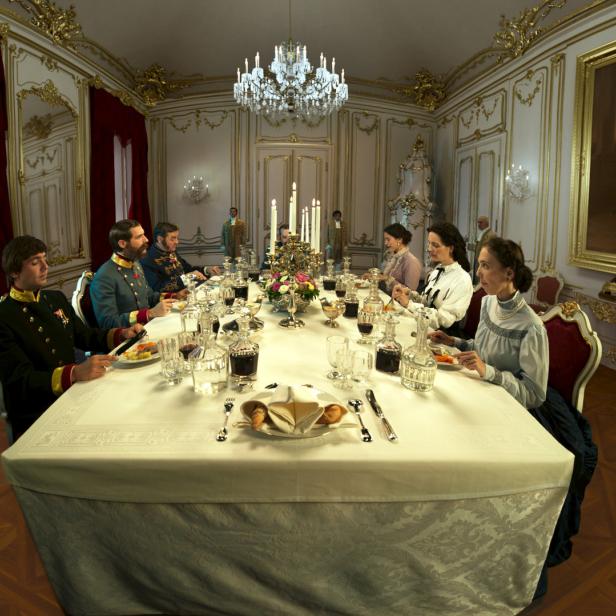 Eine Gruppe von Personen in historischen Kostümen sitzt an einem reich gedeckten Tisch in einem opulenten Speisesaal.