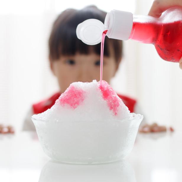 Roter Sirup wird auf einen Berg aus Schabeis gegossen, während ein Kind im Hintergrund zusieht.