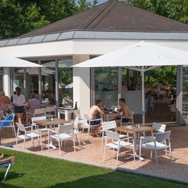 Gäste sitzen an Tischen im Freien vor einem Strandclub.