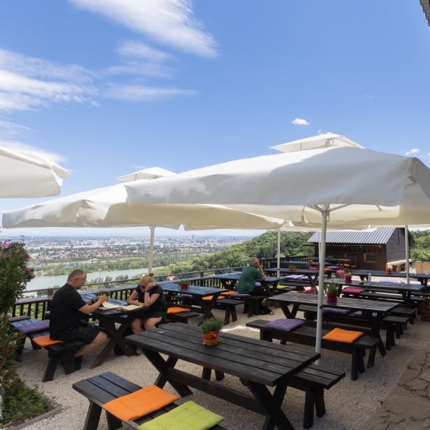 Ein Restaurant im Freien mit Tischen und Bänken unter Sonnenschirmen bietet einen Blick auf die Stadt.