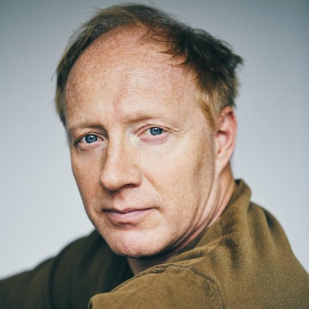 Porträt eines Mannes mit roten Haaren und blauen Augen, der einen braunen Pullover trägt.