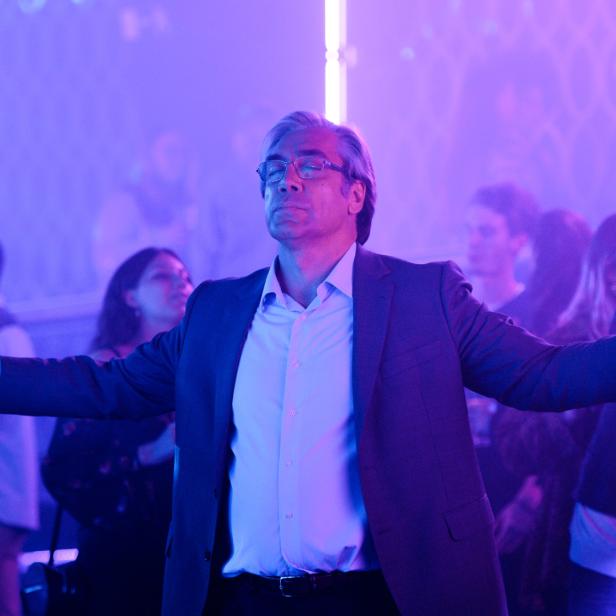Javier Bardem steht mit ausgestreckten Armen und je einem Drink in der Hand in einem Club.