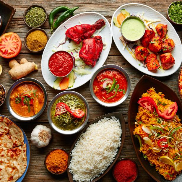 Eine Auswahl an indischen Gerichten, darunter Curry, Reis, Tandoori-Hähnchen und Naan-Brot, auf einem Holztisch angerichtet.