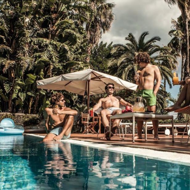 Eine Gruppe von Menschen entspannt sich mit Getränken an einem sonnigen Pool unter Palmen.