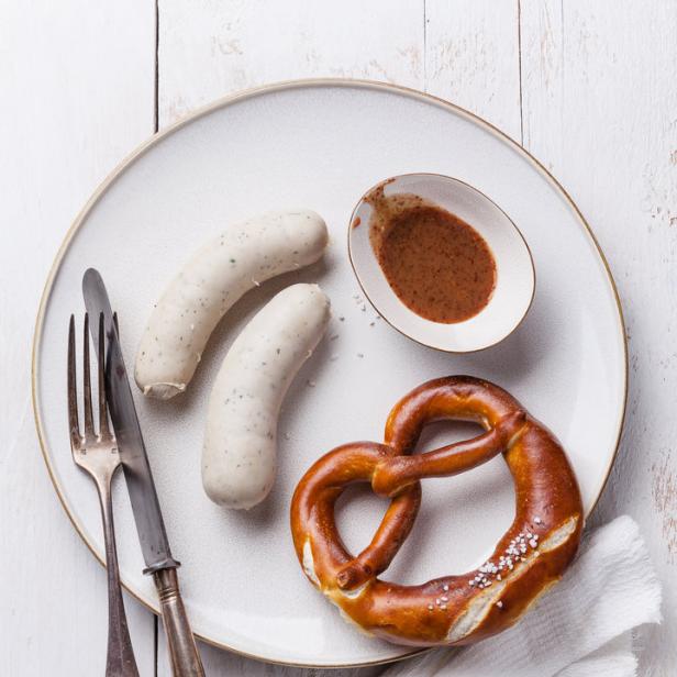 Eine Brezel, zwei Weißwürste, Messer und Gabel auf einem Teller mit süßem Senf.