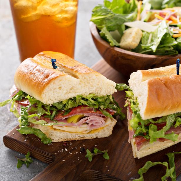 Ein aufgeschnittenes Sandwich mit Salat und Aufschnitt auf einem Holzbrett, dazu ein Salat und ein Getränk.