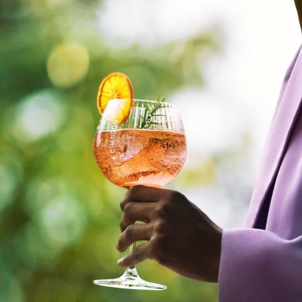 Eine Person im lila Blazer hält ein Glas Aperol Spritz mit Orangenscheibe.