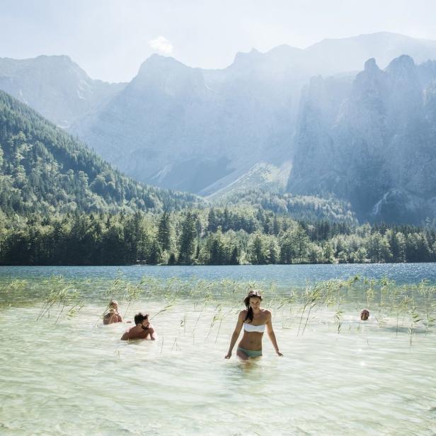 Eine Gruppe von Menschen watet in einem klaren Bergsee vor einer bewaldeten Bergkulisse.