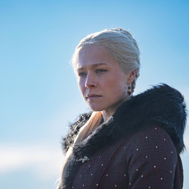 Rhaenyra Targaryen mit blonden Haaren und dunklem Umhang vor blauem Himmel.