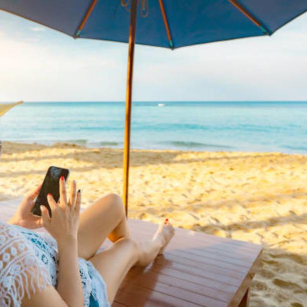 Eine Frau liegt am Strand unter einem Sonnenschirm und benutzt ihr Smartphone.