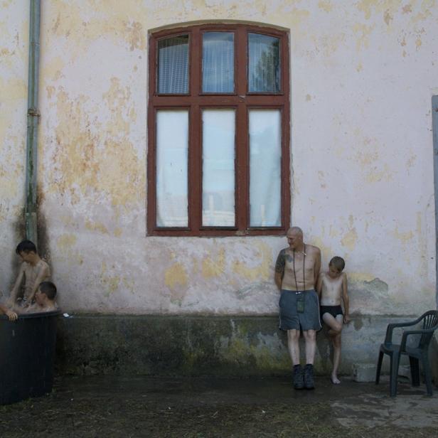 Eine Gruppe Kinder badet in einem Bottich vor einem Haus, während ein Mann zusieht.