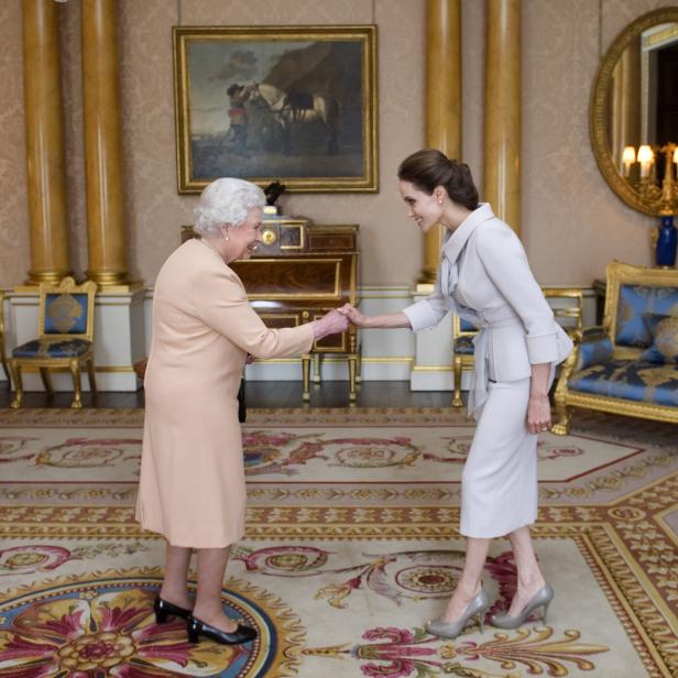 Königin Elisabeth II. und Angelina Jolie geben sich die Hand in einem opulenten Raum.