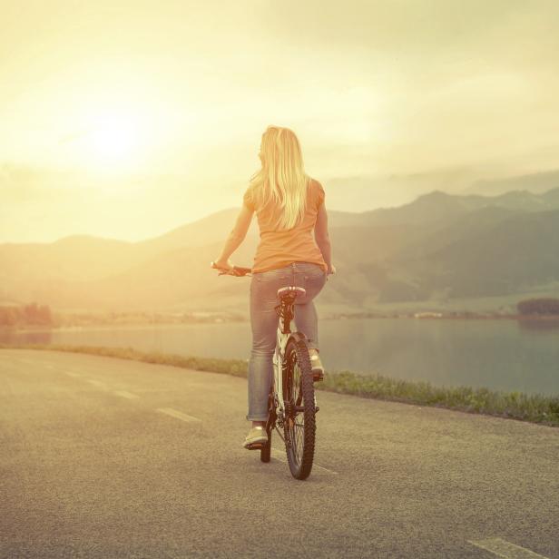 Eine Frau fährt mit dem Fahrrad auf einer Straße in Richtung eines Sees und der Berge.